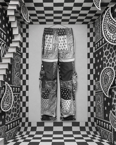 Il cargo Romanticbandana, patchwork di bandane nere, bianche e grigie, appeso con un filo a un chiodo sulla parete di fondo di una stanza di carta a scacchi bianchi e neri. A sinistra una scala bianca, sulle pareti paisley, stelle, zebrato, maculato e piccoli teschi.